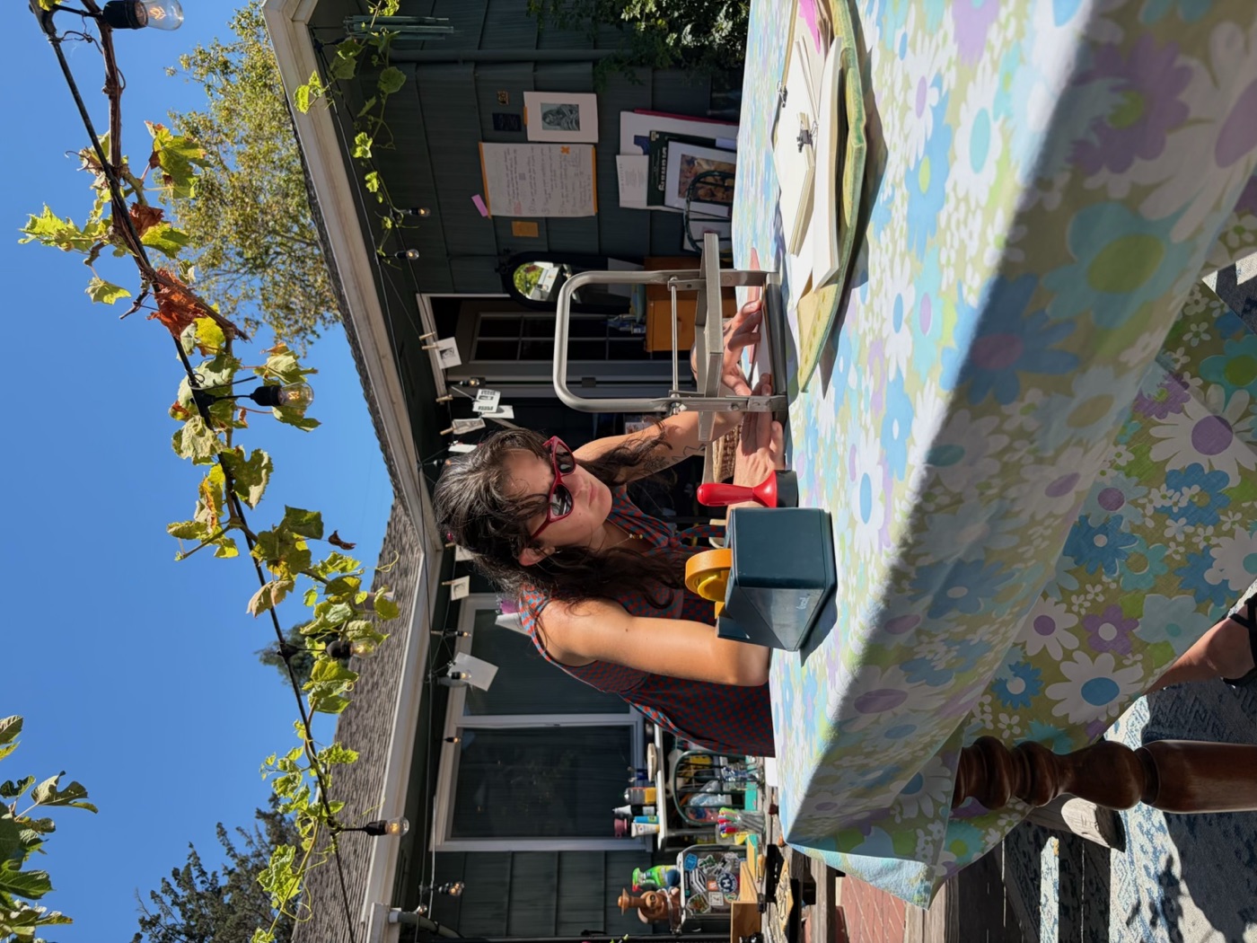 A person leans over a table of art supplies, working outdoors with plants and a dark building behind.