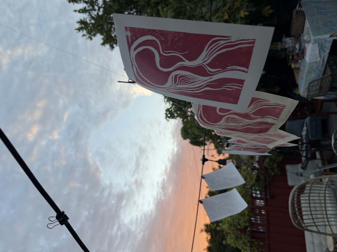 Identical pink linocut prints hang from clotheslines at sunset with trees in the background.