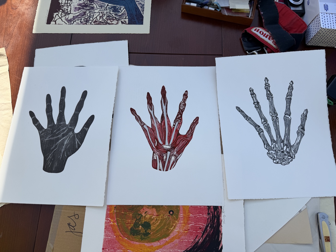 Three sheets of hand anatomy studies: line drawing, red ink, and black ink, on a wooden surface.