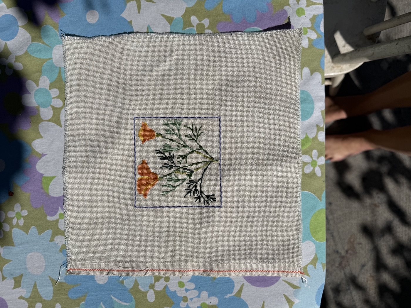 A small cross-stitch of two rust-coloured flowers with green foliage on cream linen.