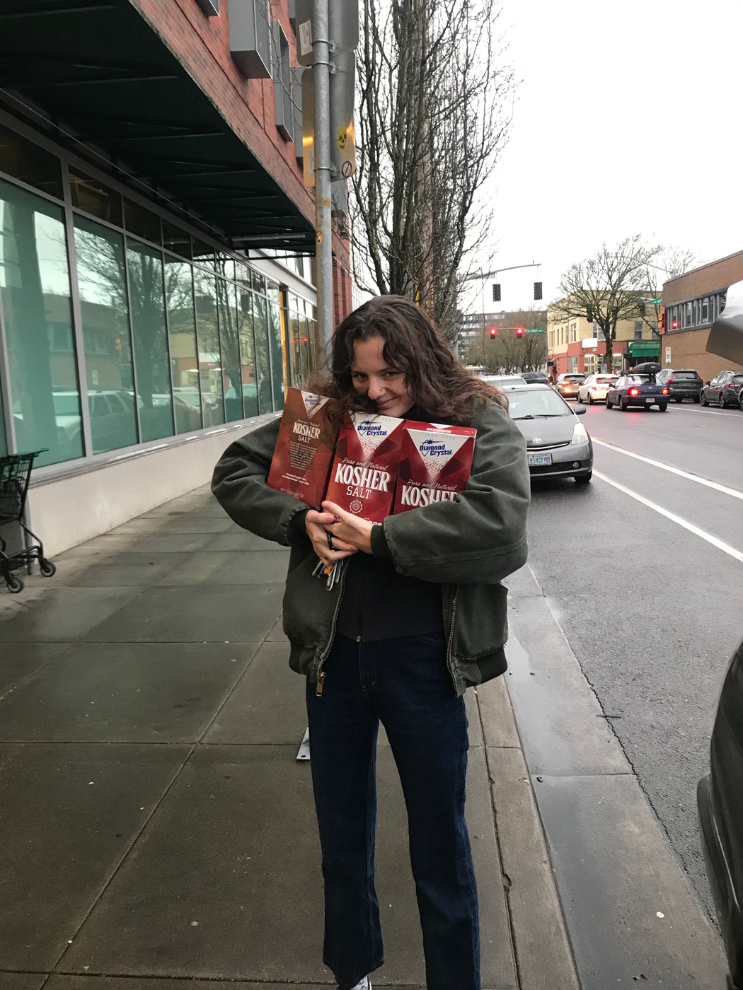 A person on a city sidewalk holding several boxes of kosher salt, storefronts and parked cars behind.