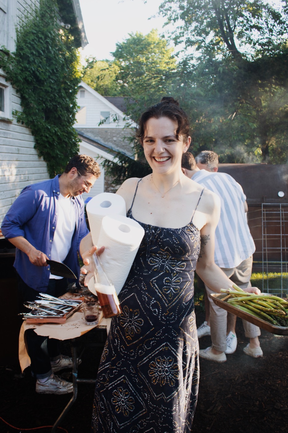 Three people at an outdoor table, the middle one holding paper towels while prepping green vegetables.