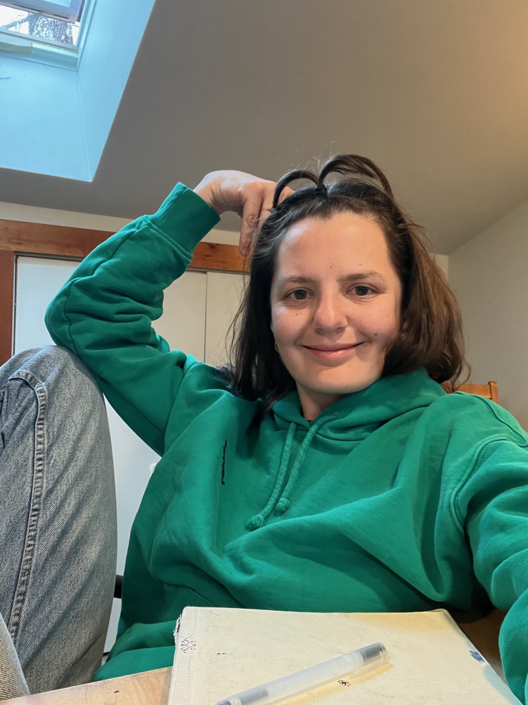 A person in a green hoodie at a table with art materials and a workspace setup.